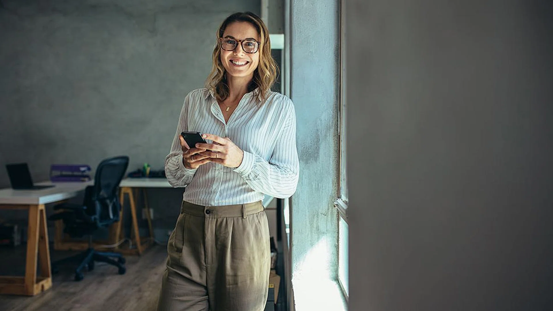 Smiling woman in casuals standing in office. Businesswoman with mobile phone in hand looking at camera.