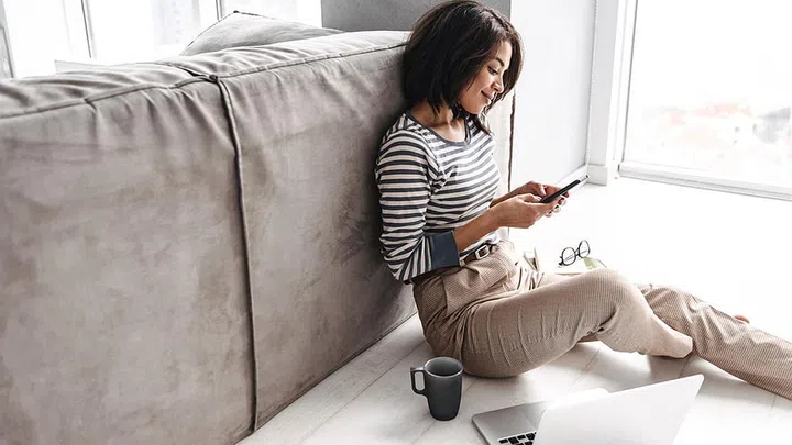 Attractive young afro american woman sitting at a couch at home, using laptop computer, drinking coffee, using mobile phone