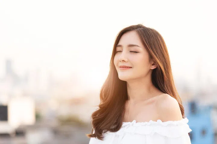 Beautiful Asian Woman is breathing fresh air. Smiling Female is standing on top of house roof over city background at outdoor. Relaxation, Eyes closed.