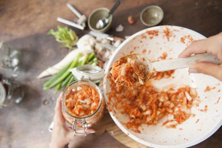 First person view of a woman scooping the vegetable/spice mixture into a large swivel top jar. She is preparing a healthy fermented, originally Korean, side dish: kimchi.