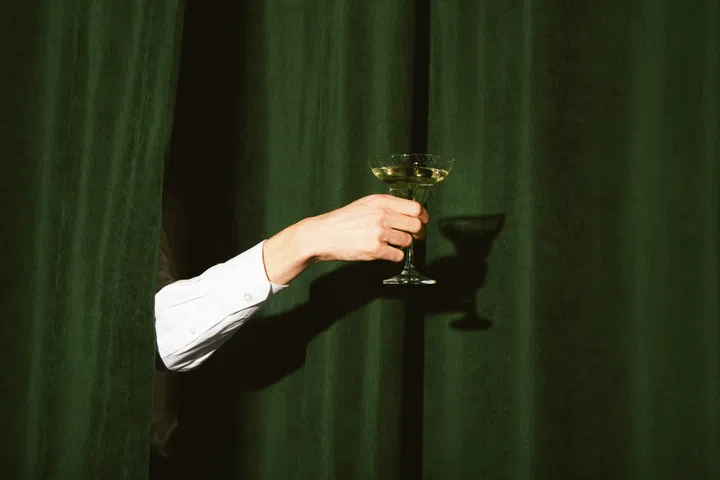 Hand with a martini glass sticks out from the green curtains