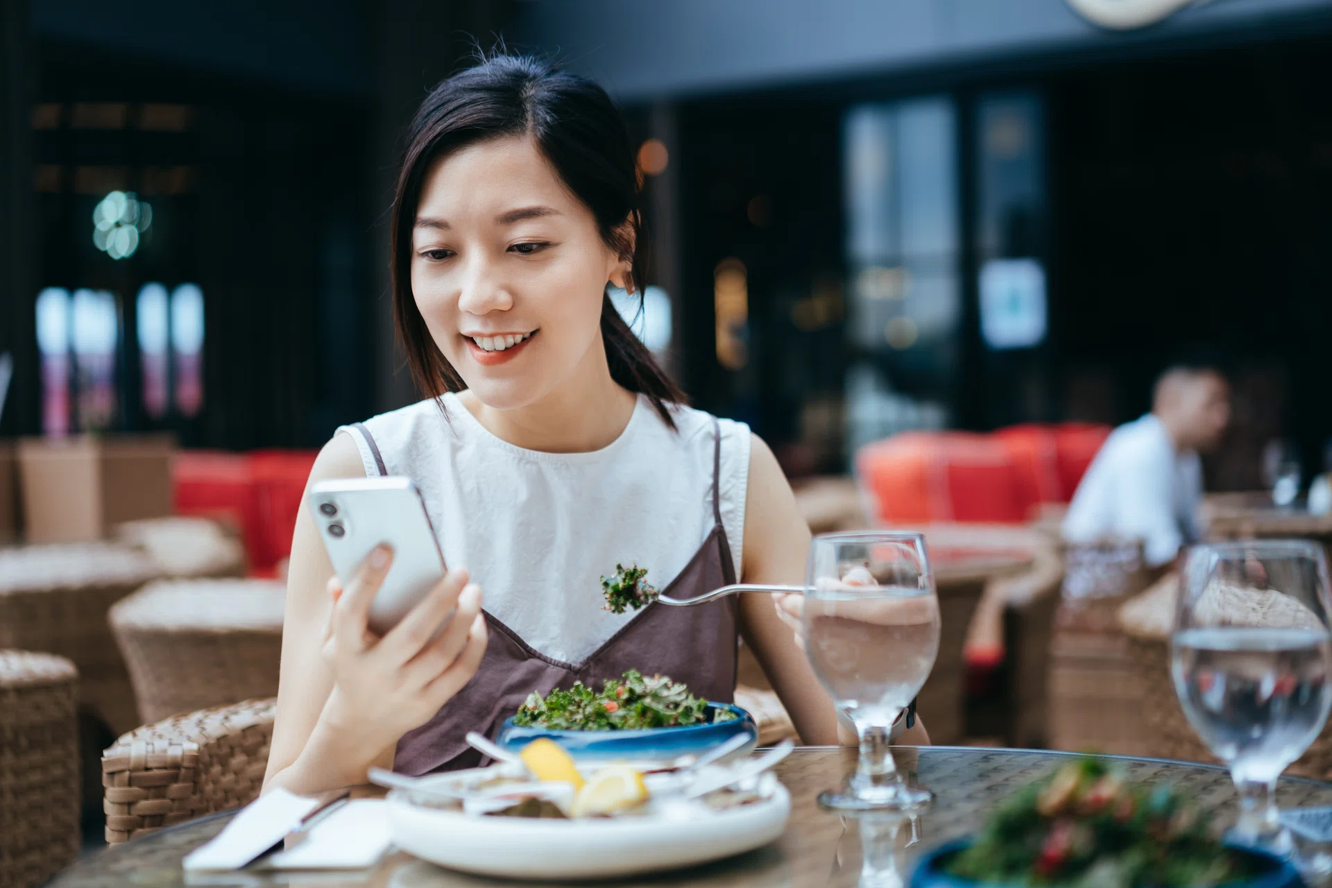 Beautiful smiling young Asian woman enjoying a bowl of healthy fresh green salad in outdoor restaurant, using smartphone during meal. Enjoying a relaxing time in the afternoon. Eating out lifestyle. Lifestyle and technology. Outdoor dining concept