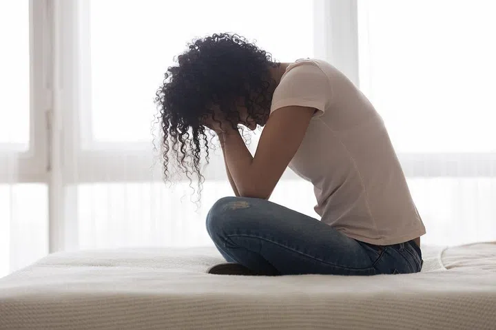 Upset sad African American woman suffering from loneliness side view, sitting in bed alone, holding head in hands, upset girl has psychological troubles, trauma, thinking about problem in bedroom