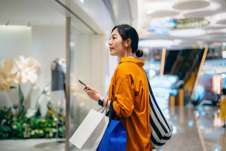 Young Asian woman with smartphone carrying paper shopping bags, standing outside a boutique looking at clothes through window in a shopping mall. Window shopping. Digital shopping technology. Sales season. Seasonal shopping and consumerism concept