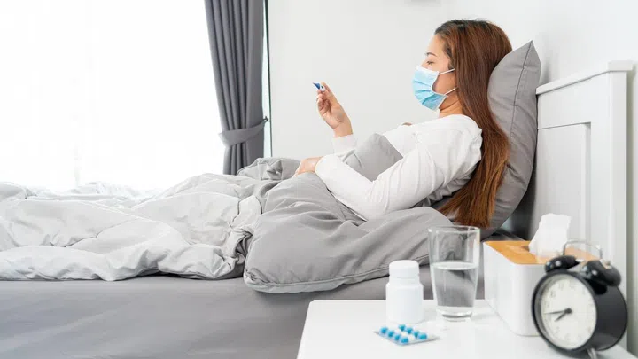 Sick Asian woman with protective mask lying in bed with high fever and measuring body temperature by thermometer and touching her forehead at her home