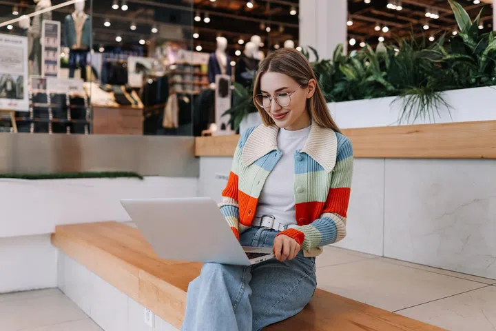 A young female freelancer influencer in glasses and fashionable casual clothes works online remotely using a laptop while sitting on the stairs in a shopping mall in a public place.