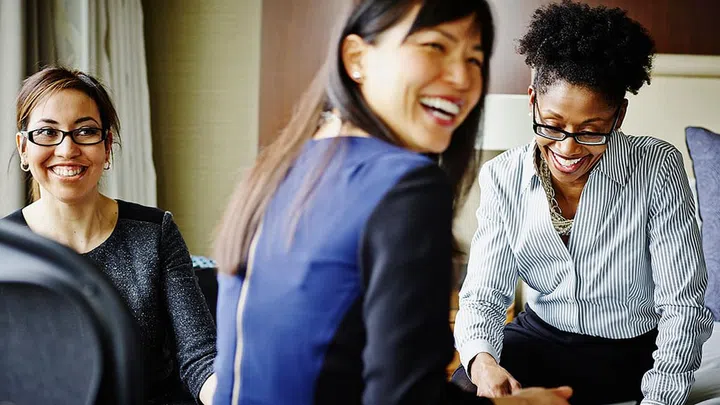 Three smiling businesswomen sitting in hotel suite discussing project details on digital tablet