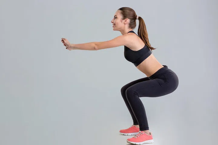 Attractive young sportswoman in sportwear doing squats isolated over grey background