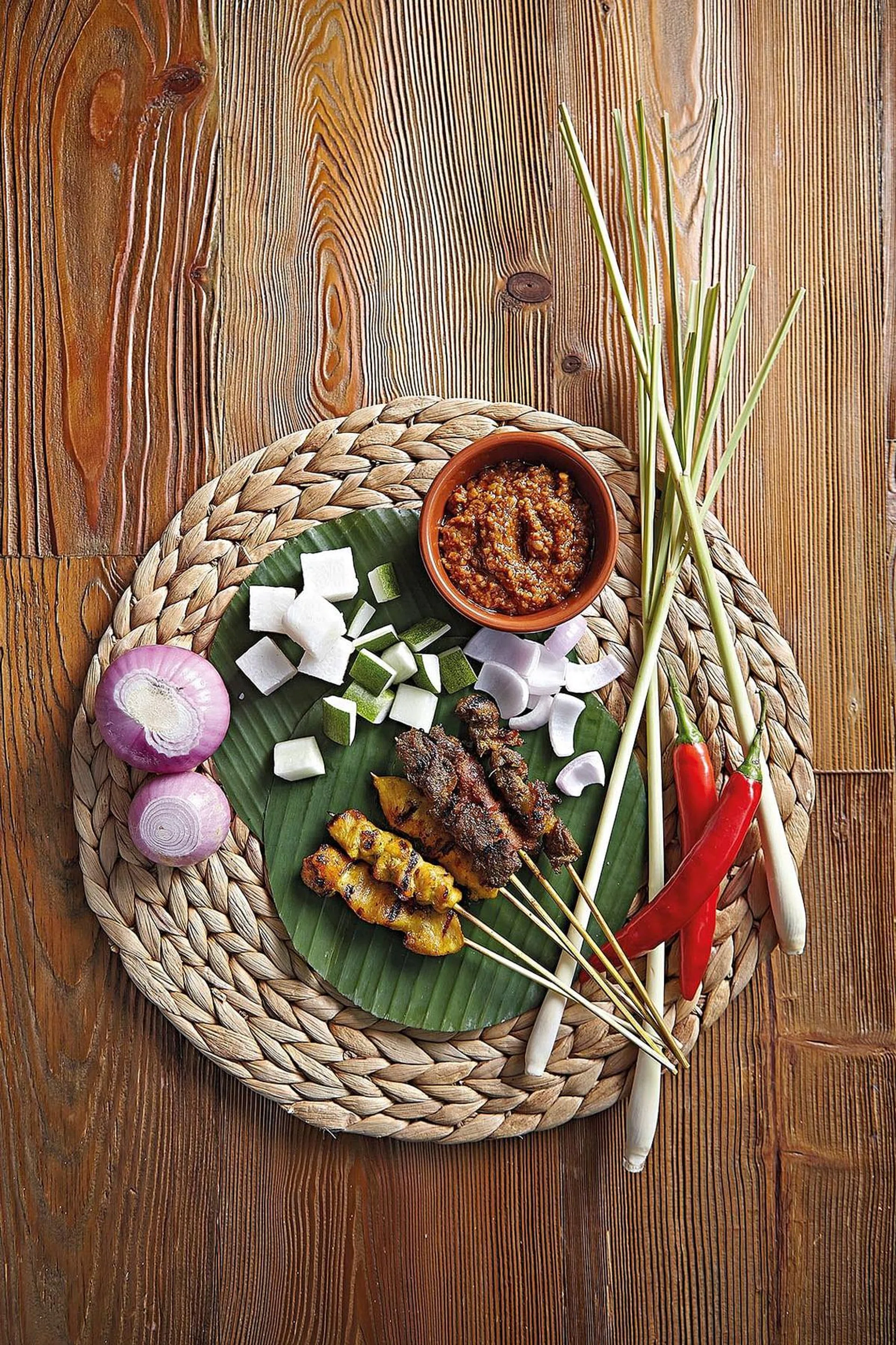 A plate of satay with stylised ingredients on a rattan straw mat.