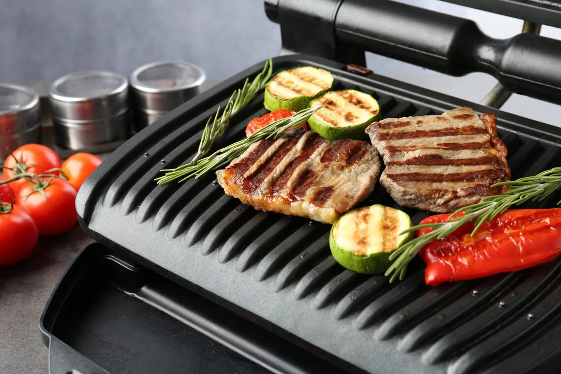 Electric grill with beef steaks, vegetables and rosemary on gray table, closeup
