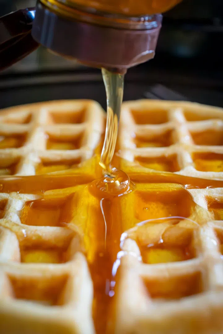 Locally sourced raw honey is added from a container dripping onto a waffle below.