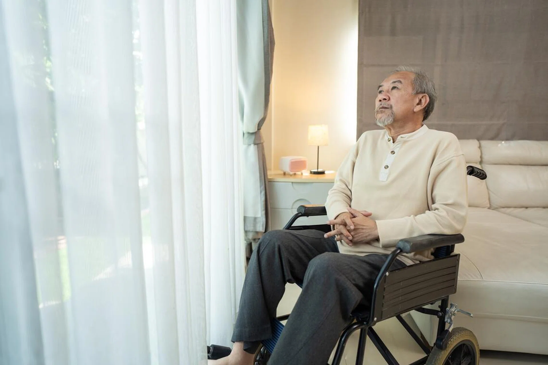 Asian senior elderly male sitting on wheelchair in living room at home. Unhappy older man feeling lonely and worry about health care problem after retirement. Hospital Healthcare and medical concept.