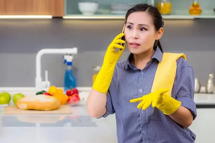 A domestic helper in uniform and yellow cleaning gloves in a kitchen talking on the phone. Photo 123RF