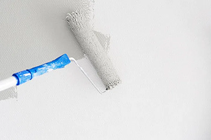 A white roller paint brush with a blue handle painting a wall white.