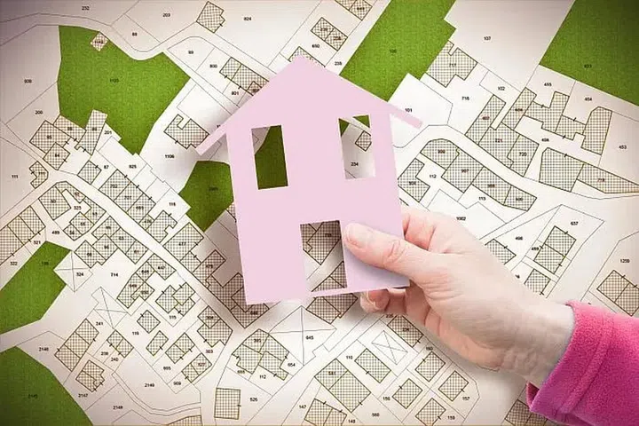 Hand holding a pink paper cut-out of a house against a map. Photo 123RF