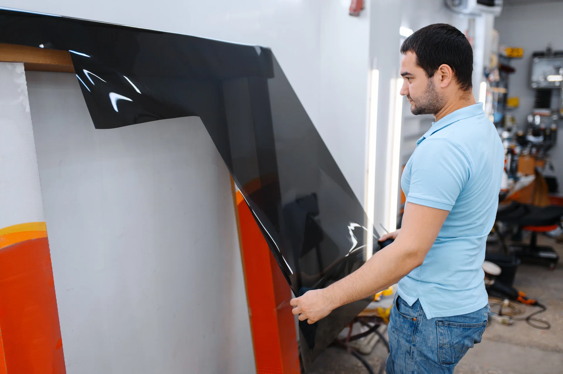 Male worker holds roll of car tinting, tuning service. Mechanic applying vinyl tint on vehicle window in garage, tinted automobile glass.
