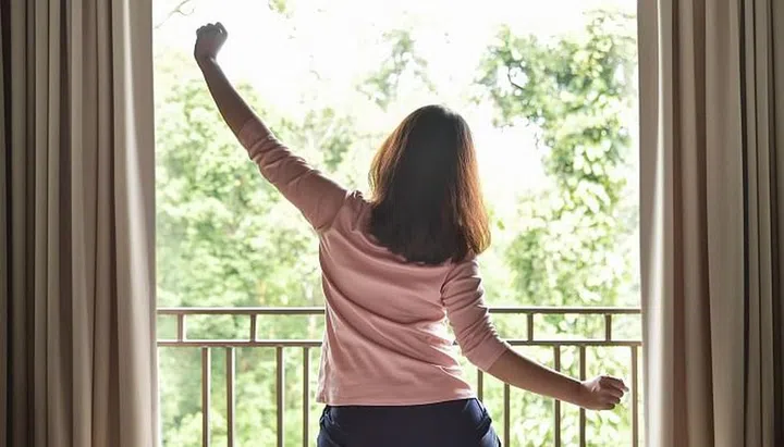 Rear view. Happy, Relaxed Asian woman waking up and Outstretched her arms on green natural background at her bedroom in the morning. Resting on Holiday.