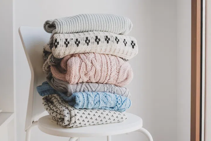 Pile of warm sweaters on white chair. Woolen knitwear clothes for autumn and winter season. Handmade jumpers.