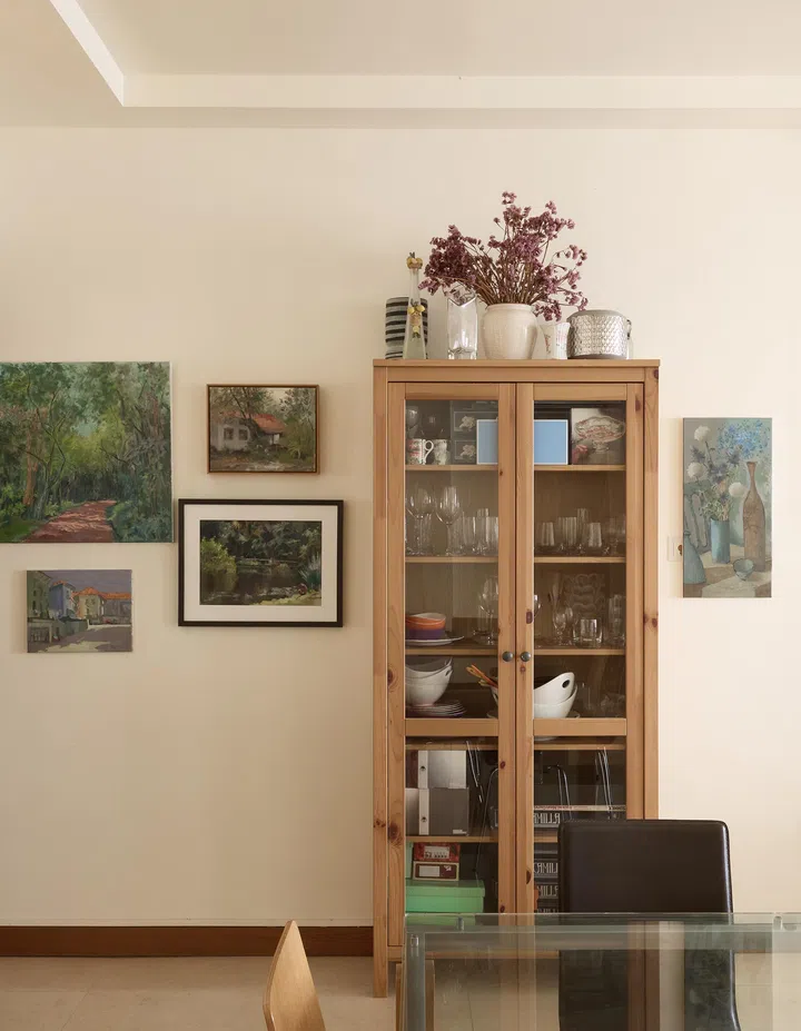 A glass-front cabinet flanked by small paintings and filled with decorative items and collectibles.
