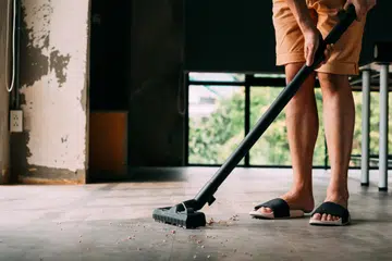 Low section of human legs wearing sandals using industrial electronic vacuum cleaner to hoover inside the house with plenty of dust and dirt - cleanliness and housekeeping concept