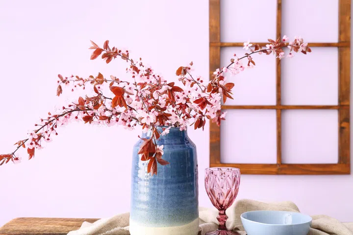 Vase with blooming tree branches, glass, bowl and napkin on wooden table near pink wall.