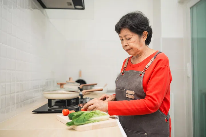 Elderly woman preparing fresh vegetables in modern kitchen, chopping tomatoes and lettuce, focus on healthy cooking and culinary skills at home.