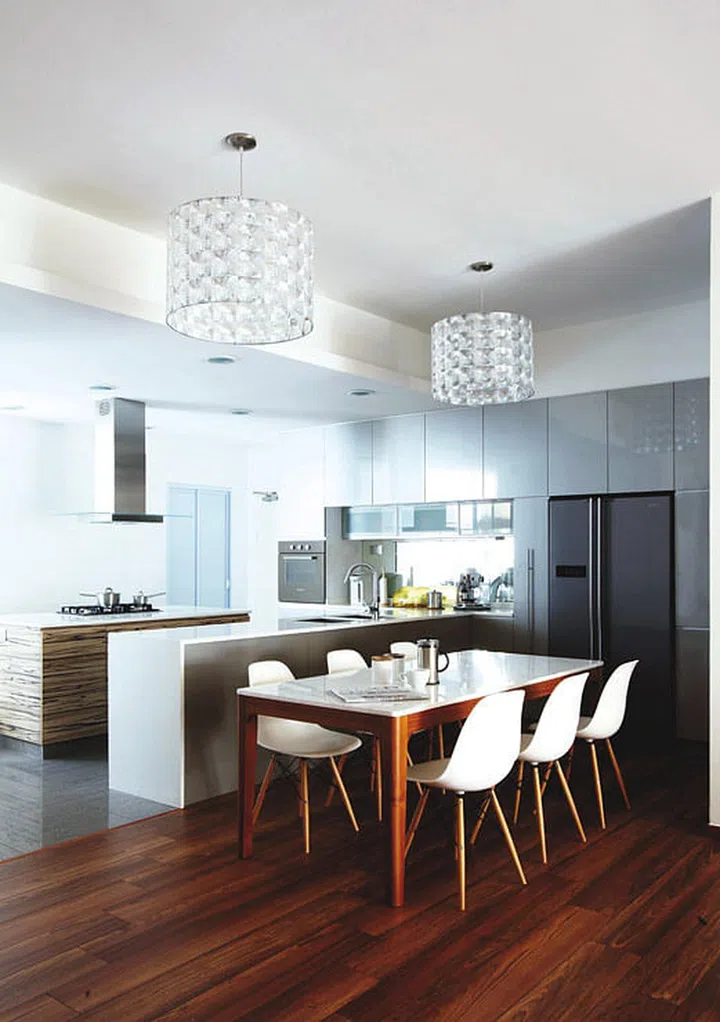 The cool white and grey tones are balaced by warm wood hues for a palette that never goes out of fashion. The high-gloss greys of the tiled flooring and Formica laminate on the cabinets create a chic contrast with the matte wood tones of the dining area and cooking island.