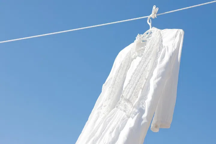 Washed white dress hanging outdoors on a laundry line against a blue sky background.