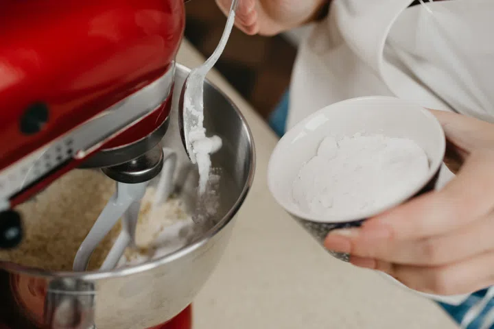 A close photo of powdered sugar is falling from the spoon into the stainless steel bowl of the red stand mixer which is mixing shortcrust pastry.
