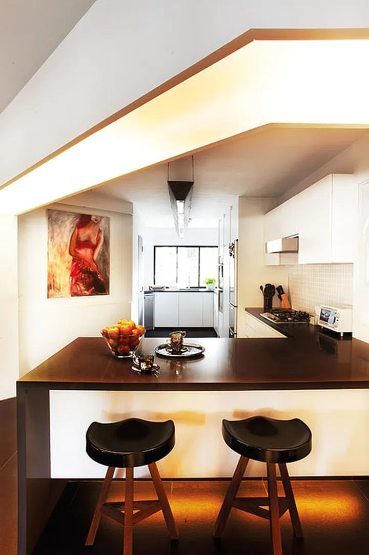 The wall separating the kitchen from the living room was torn down and the wet area moved to the far end of the kitchen/ utility area. The L-shaped counter runs on the side, incorporating the gas hob and hood for cooking, and doubles up as a dining table, too. A custom-made suspended lamp made of brushed steel contrasts with the sleek appliances.