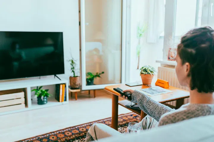Teenage girl in casual clothes, sitting on comfortable sofa in apartment, holding glass of water in hands, watching TV. Far viewing angle. soft focus