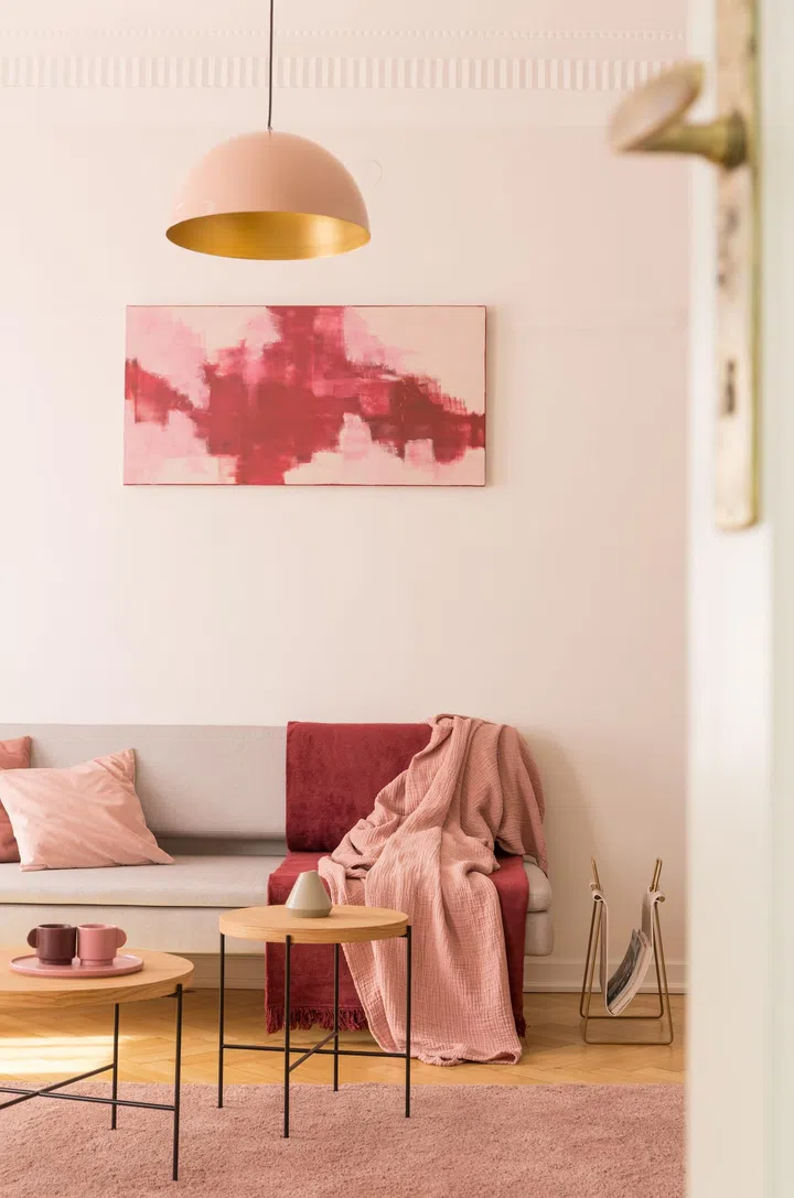 Lamp above wooden tables on pink carpet in loft interior with red poster above grey sofa. Real photo.