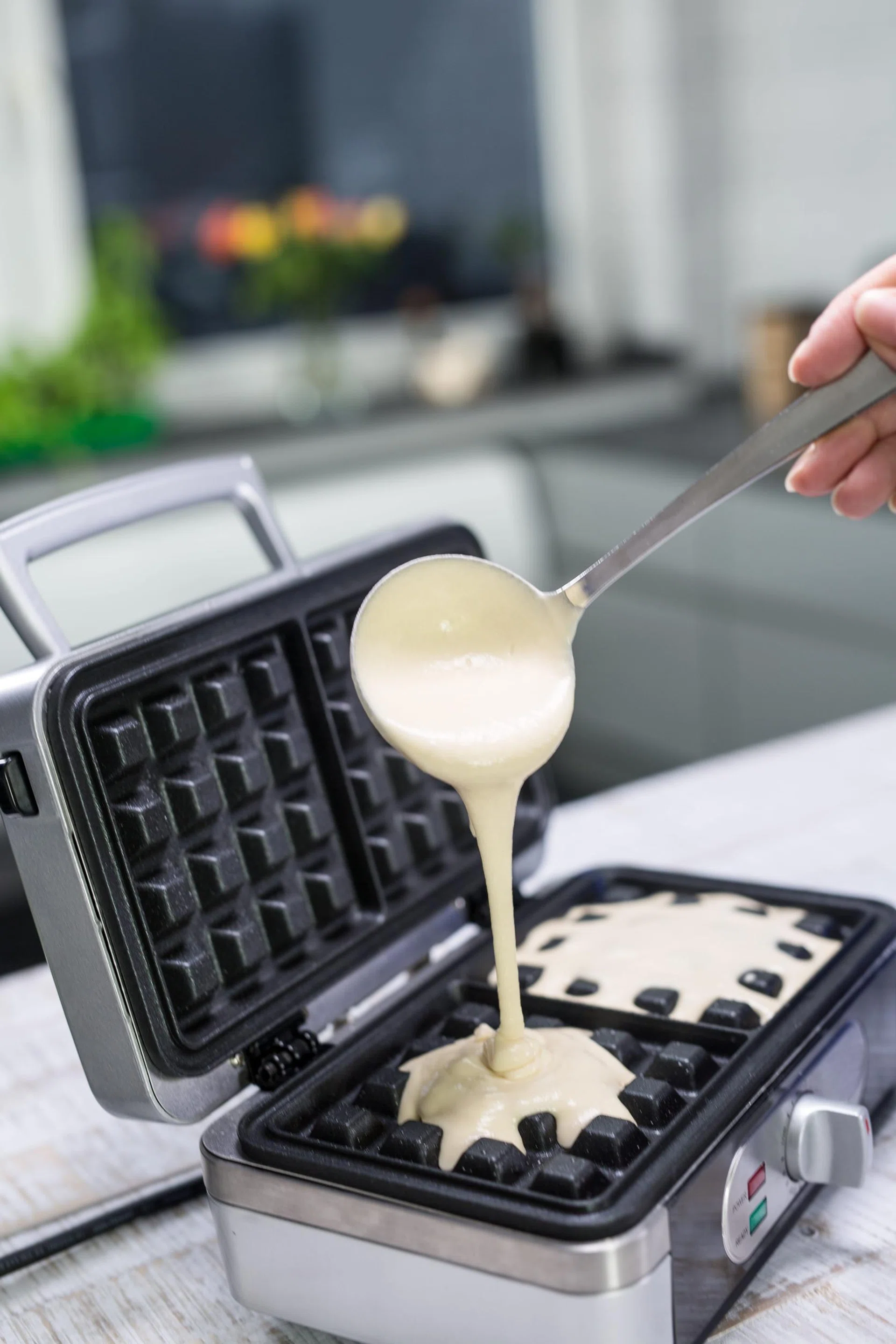 A people hand is using a ladle to pour dough on a waffle iron in a modern kitchen.