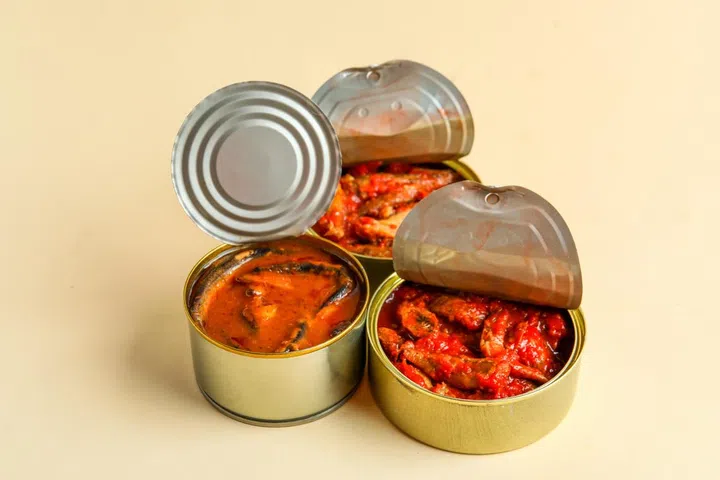 Opened tin cans with fish in tomato sauce on beige background