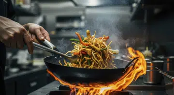 Chef hands rapidly tossing stir fried noodles and vegetables in a sizzling wok over intense flames in a professional kitchen.