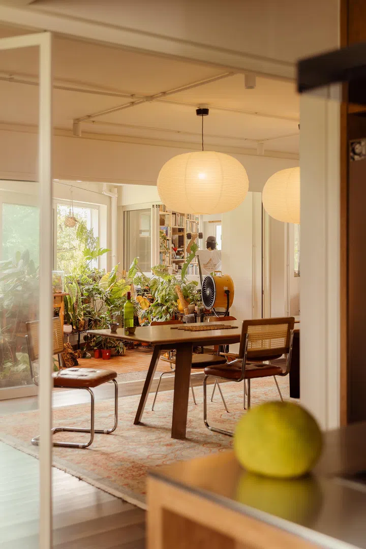 The dining area is anchored by a vintage rug and oversized paper lanterns, with greenery spilling in from the balcony and a sightline into the study.
