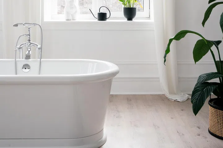 Vertical shot of modern bathroom with white freestanding bathtub against large window and green potted plants. 