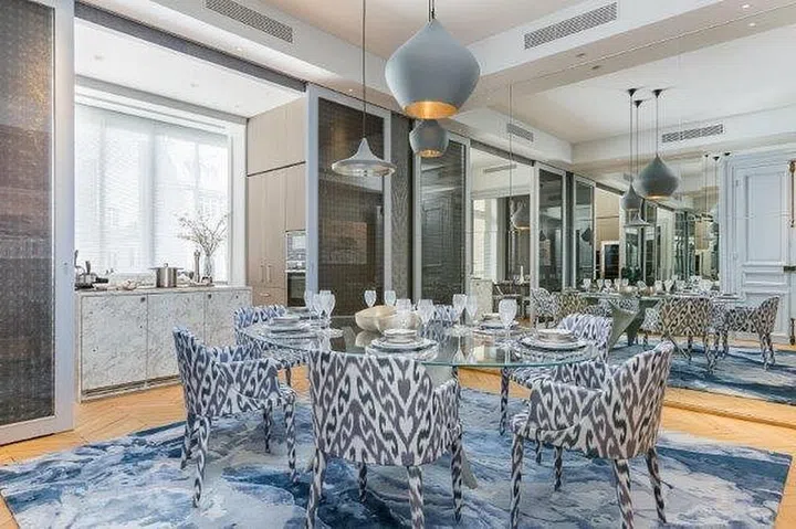 Mirrors on either side of the dining room create infinite reflections, multiplying the brightness and airiness of the space. The blue and white Sahrai Helix rug with a dreamy print stylishly clashes with the dining chairs that are upholstered in a Dedar Silkyway Bermude modern ikat printed fabric.