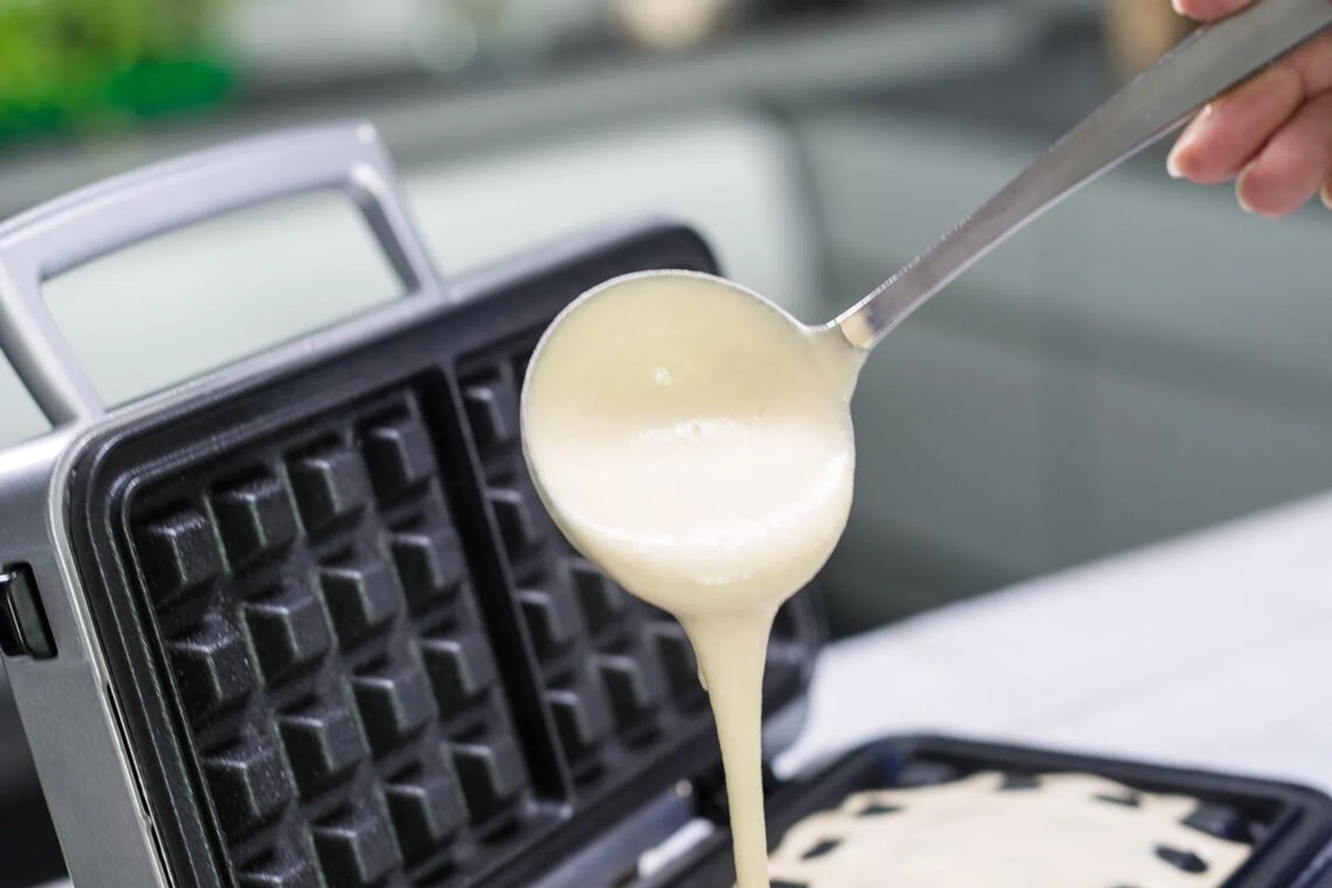 A people hand is using a ladle to pour dough on a waffle iron in a modern kitchen.