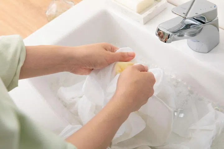 Woman's hands washing a piece of white cloth in a bubbly white basin sink.