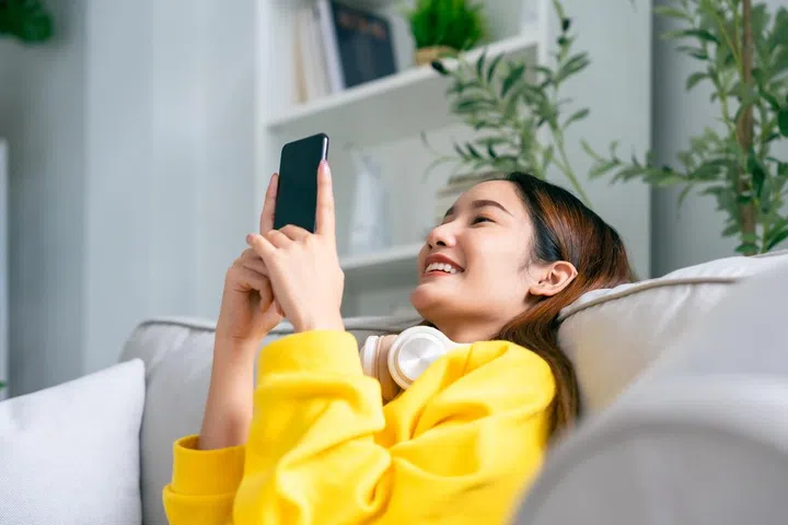 Happy young asian woman wearing headset and relaxing on comfortable couch at home, texting messaging on smartphone, smiling girl use cellphone chatting, online shopping at home, video call