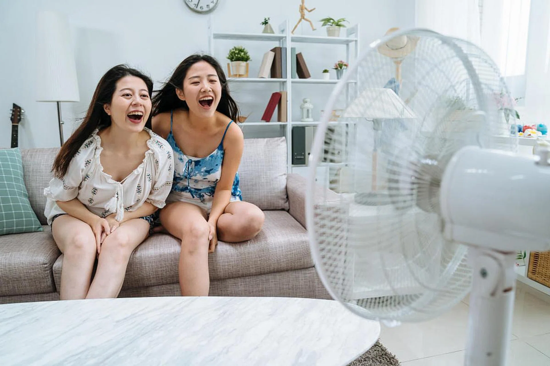 two-young-asian-girl-friends-sitting-couch-sofa-cozy-living-room-playing-with-electric-fan-playful-women-having-fun-shouting-yelling-air-cooler-machine-apartment-by-window-summer-1