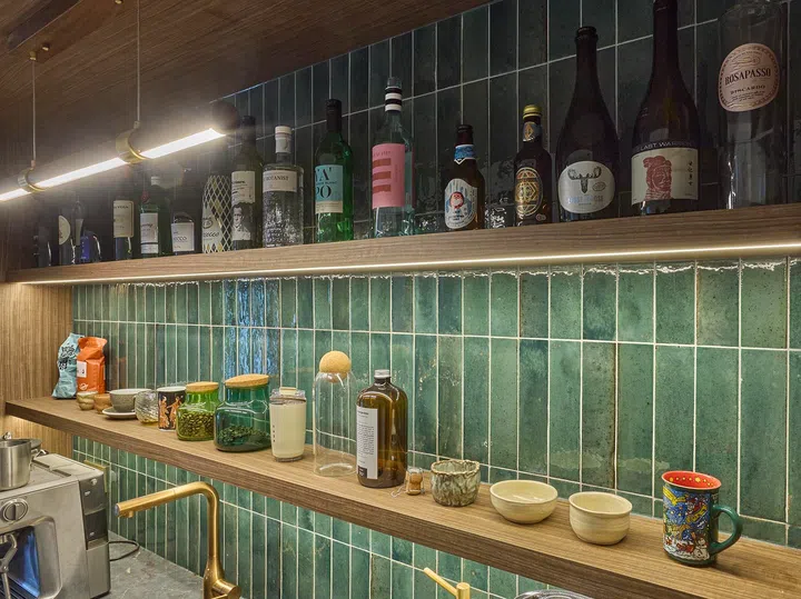 Green ceramic tiles and warm lighting set the backdrop for a collection of memorable bottles, arranged alongside glassware and everyday objects within easy reach.