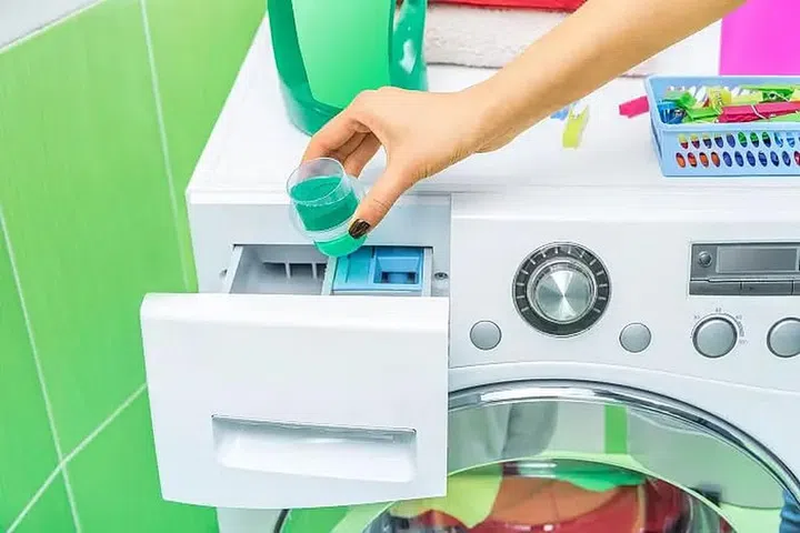 A pair of manicured female hands pouring a cup of laundry detergent into the detergent drawer of a washing machine. Photo 123RF
