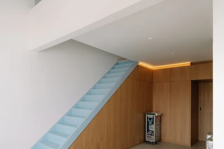 Tucked beneath the pale blue staircase, the walk-in wardrobe is lined with full-height bespoke oak cabinetry that maximises storage while maintaining the home’s restrained material palette.