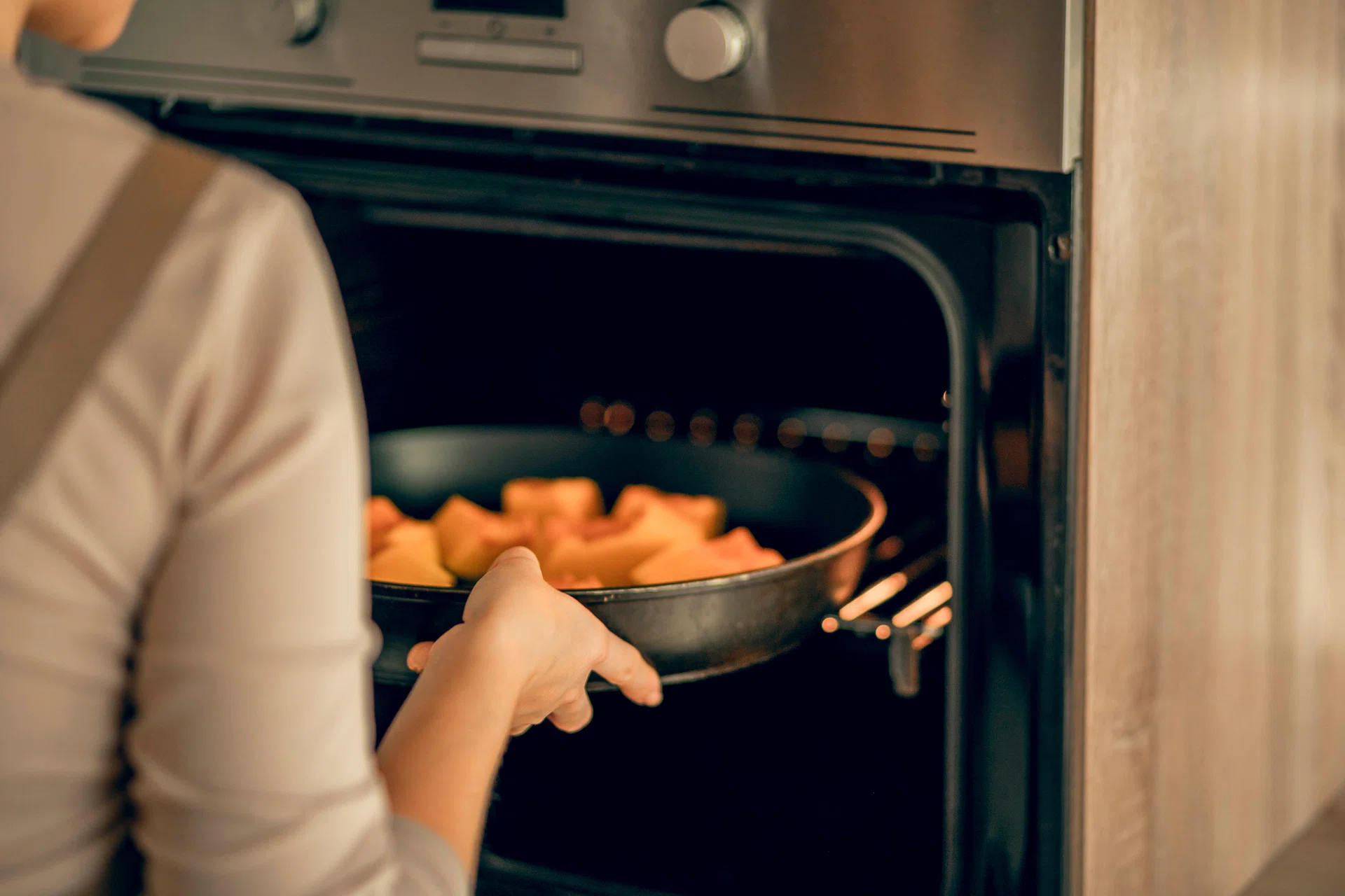 A young woman in an apron puts a dish with pumpkin in an electric oven for baking in the kitchen.