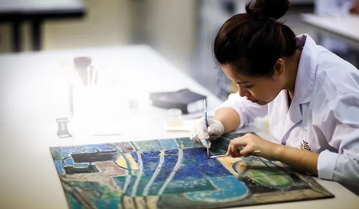A woman in white lab coat restoring a piece of artwork on a table.