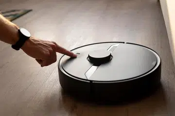 A person’s hand pressing a button on a robot vacuum cleaner on the floor.