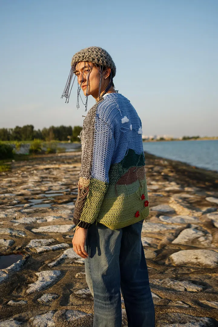 Edwin Goh站在海边防波堤上，回头望向镜头，身穿钩织作品《Storm Sweatshirt》，并搭配《Raincloud Beanie》针织帽。毛衣以灰、蓝、绿色块拼接而成，背面织有白色云朵、山丘及红色花朵等元素，并刻意保留部分垂落的线头，象征风暴过后逐渐重现希望与生命力。针织帽饰有垂坠流苏，呼应雨丝的意象。夕阳洒落在人物与作品上，远方是海岸线、树林与辽阔天空，营造宁静而充满希望的氛围，呼应作品关于走过低潮、迎向重新开始的主题。