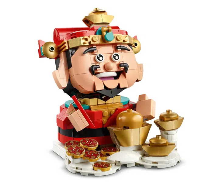 画面展示一款 LEGO 财神爷（Fortune Master，40864）积木摆设模型。主体为一位笑脸财神造型人偶，头戴红色与金色装饰的传统头冠，脸部表情夸张可爱，带有胡须与圆润五官。人物身穿红色服饰，手持红色配件，整体配色以红、金与肤色积木为主。底座前方摆放多枚金色与红色图案的金币积木，以及多层堆叠的金元宝造型摆件，营造出富足与喜庆的视觉效果。整体造型立体、色彩鲜明，呈现传统新春节庆中象征财富与祝福的财神形象，适合作为居家空间的新春展示摆设或收藏装饰。